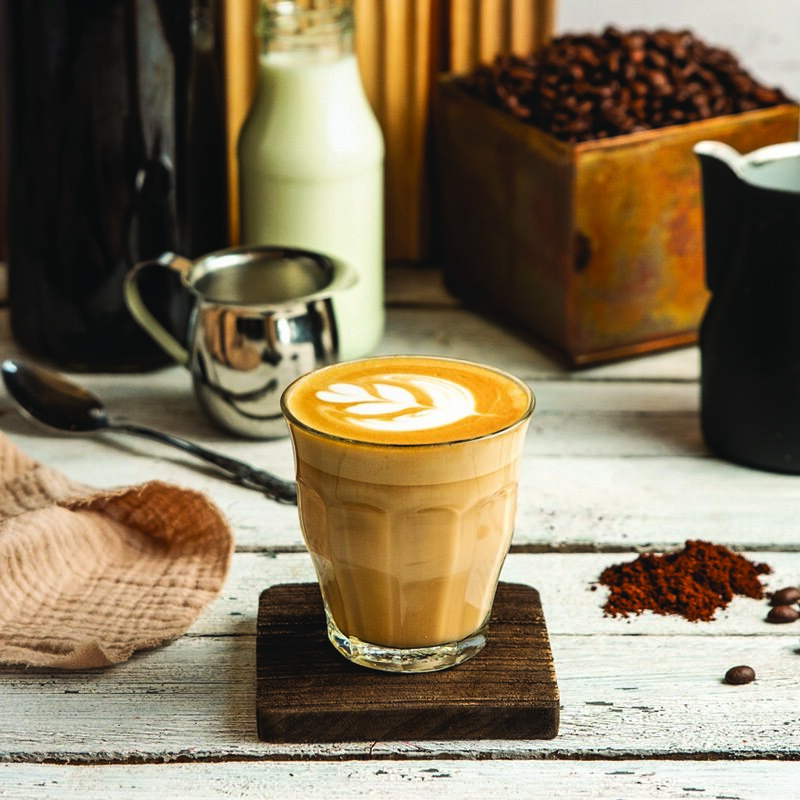 .כוס קפה לאטה עם ציור לב בקצף, מונחת על תחתית עץ, ברקע קופסת פולי קפה, בקבוקי חלב וכלי מתכת