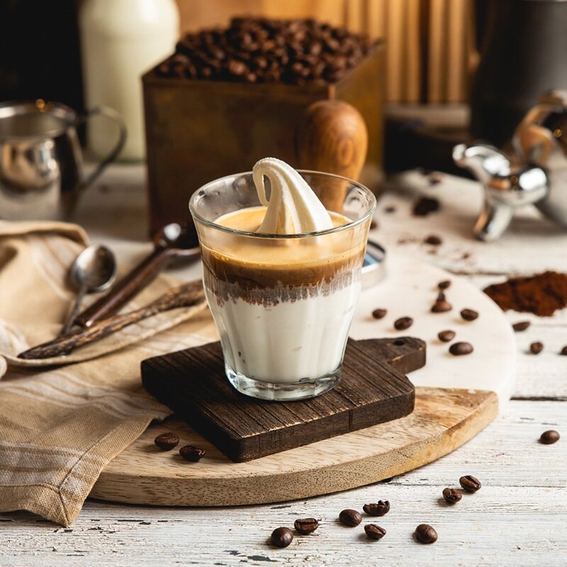.כוס זכוכית עם גלידת וניל וקפה חם מעליה, מונחת על קרש עץ קטן, ברקע פולי קפה, כפיות ובקבוק חלב