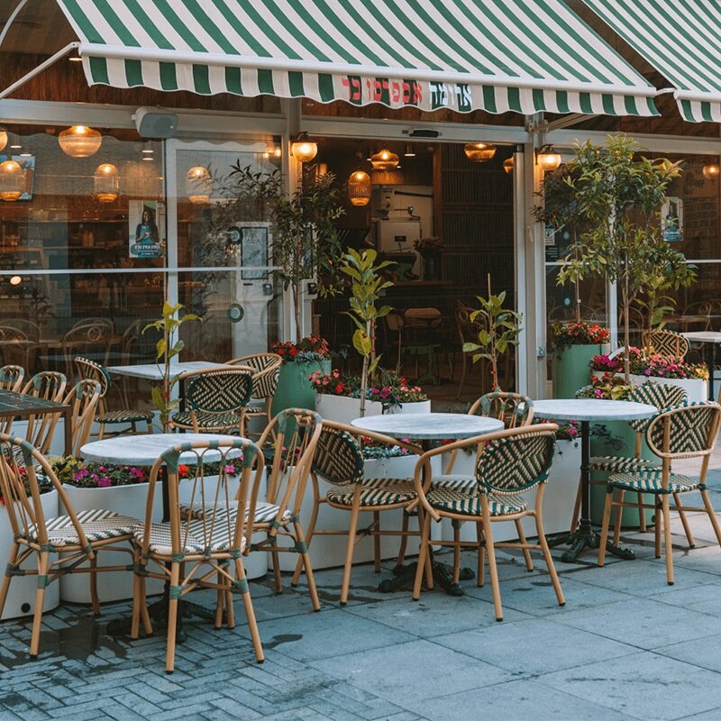 .מראה חזית של קפה ארומה אספרסו בר – שולחנות וכיסאות ישיבה בחוץ תחת סוכך פסים ירוק־לבן, עם עציצים ופרחים צבעוניים