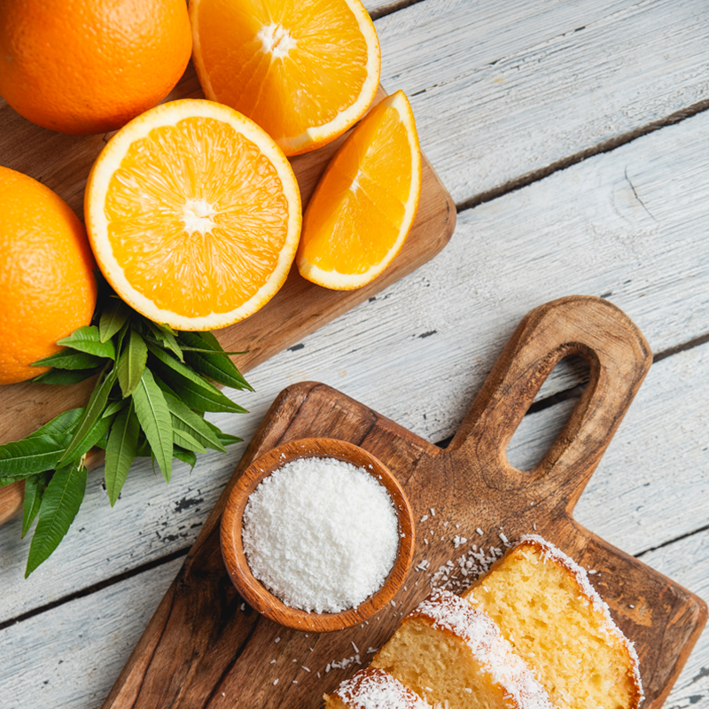 .חצאי תפוזים טריים וקערית קוקוס מגורר על קרש חיתוך, יחד עם קצה פרוסות עוגת תפוז וקוקוס