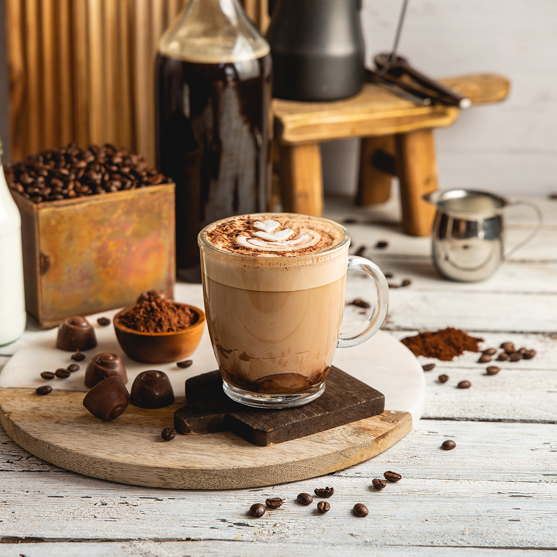.כוס קפה מוקה עם קצף חלב מעוטר בלב, מונחת על קרש עץ לצד קוביות שוקולד, אבקת קקאו ופולי קפה, עם כלי קפה ברקע
