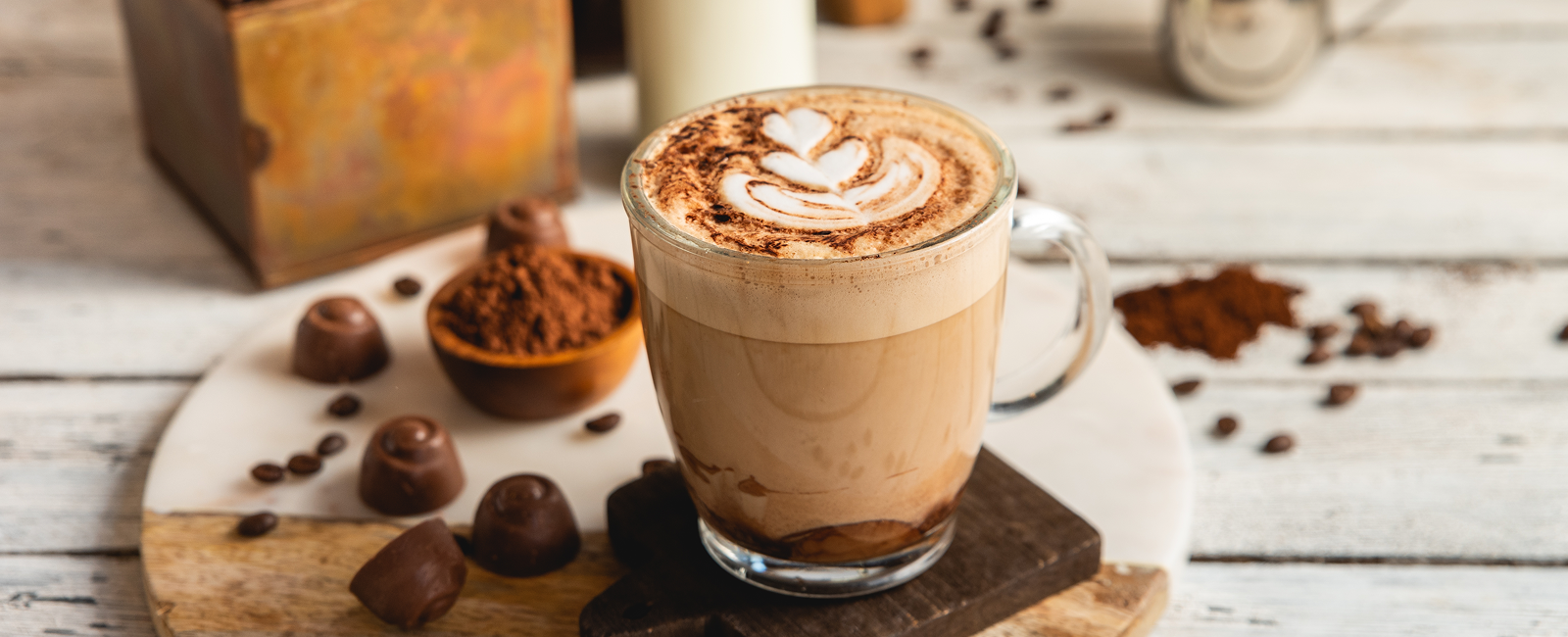 .תקריב על כוס קפה מוקה עם ציור לב בקצף, ברקע מטושטש נראים אבקת קקאו ושוקולד