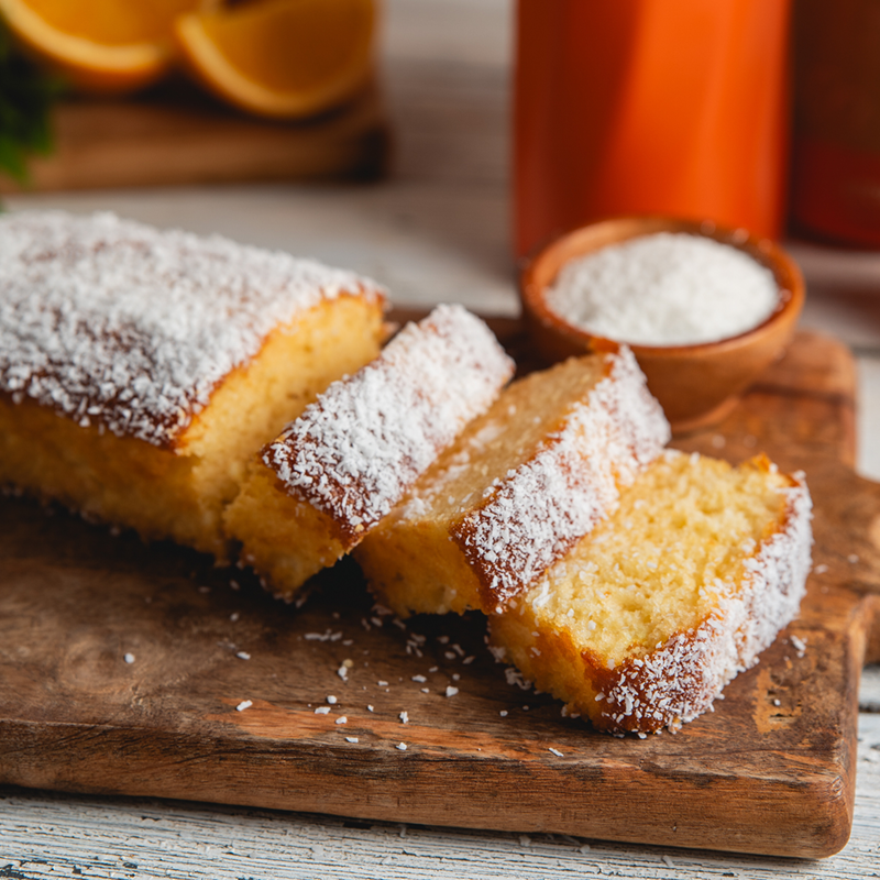 .עוגת תפוז וקוקוס פרוסה על קרש עץ, מצופה קוקוס מגורר ולצידה קערית קטנה עם קוקוס