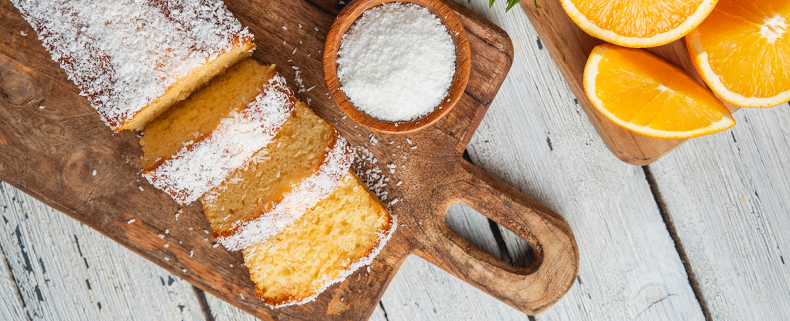 .עוגת תפוז וקוקוס פרוסה על קרש עץ עם קערית קוקוס ומצידה פלחי תפוז טריים